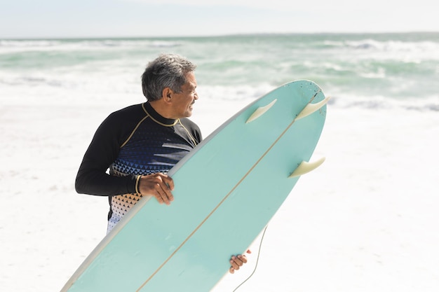 Homem sênior birracial carregando prancha azul na costa enquanto olha para o mar durante o dia ensolarado