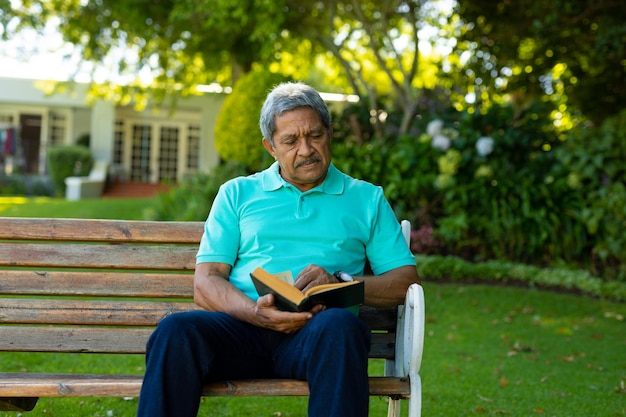 Homem sênior birracial focado lendo livro enquanto está sentado no banco contra árvores e plantas no parque