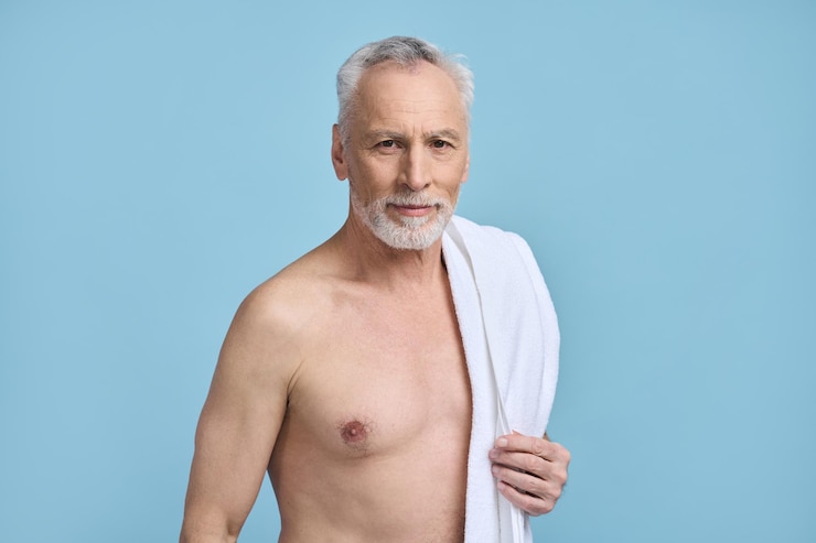 Foto homem sênior bonito e caucasiano 6069 anos de idade, segurando uma toalha branca, olhando com confiança para a câmera em pé com o tronco nu, isolado no fundo azul rotina diária beleza spa higiene