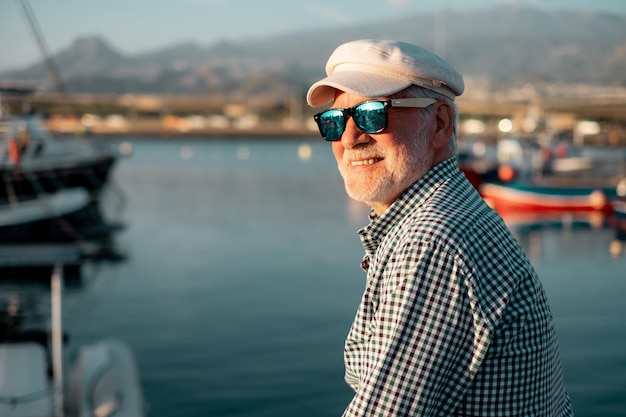 Homem sênior bonito em camisa casual e chapéu sentado no porto olhando para barcos e mar Idoso relaxado homem barbudo usando óculos escuros aproveitando a viagem ou a aposentadoria