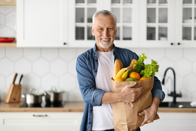 Homem sênior bonito posando na cozinha com saco de papel de mantimentos