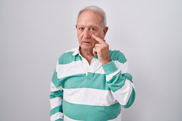 Homem sênior com cabelos grisalhos em pé sobre fundo branco apontando para o olho observando você gesticular expressão suspeita