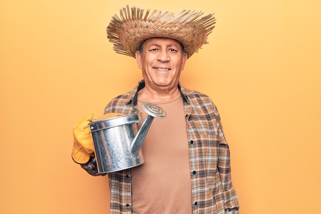 Homem sênior com cabelos grisalhos usando chapéu de jardineiro segurando regador parecendo positivo e feliz em pé e sorrindo com um sorriso confiante mostrando os dentes