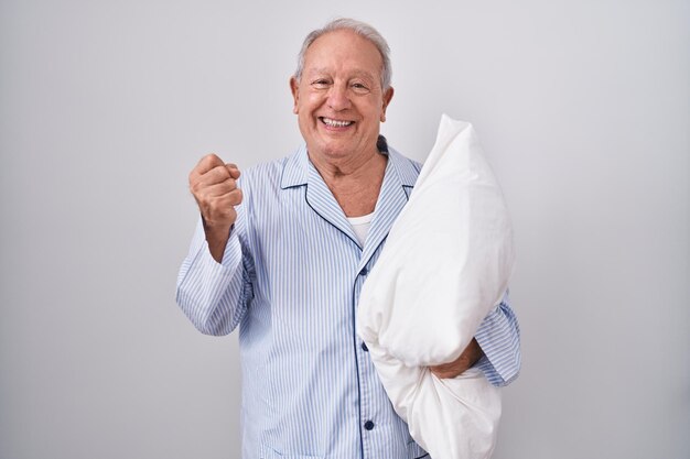 Homem sênior com cabelos grisalhos usando pijama abraçando travesseiro muito feliz e animado fazendo gesto de vencedor com os braços levantados sorrindo e gritando pelo conceito de celebração de sucesso