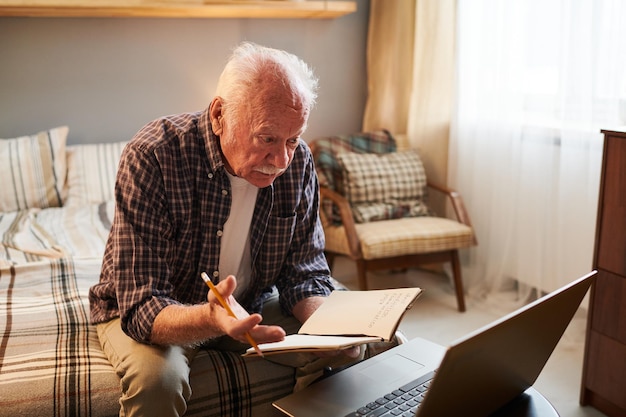 Homem sênior com caderno e lápis sentado na frente do laptop