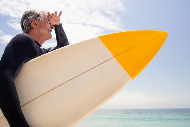 Homem sênior com prancha de surf, protegendo os olhos na praia