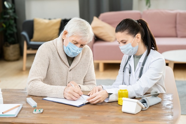 Homem sênior doente com máscara e roupa casual assinando documento médico enquanto está sentado à mesa em frente a uma jovem médica na sala de estar