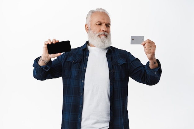 Homem sênior duvidoso e cético, mostrando a tela horizontal do smartphone e olhando com descrença no cartão de crédito, tendo dúvidas de estar inseguro sobre fundo branco