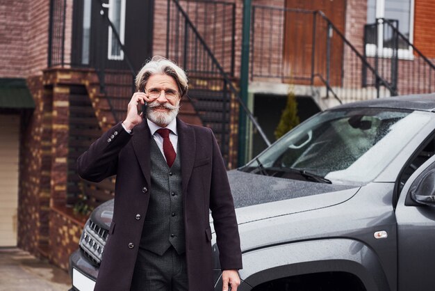 Homem sênior elegante com cabelos grisalhos e barba está ao ar livre na rua perto de seu carro ter uma conversa ao telefone.