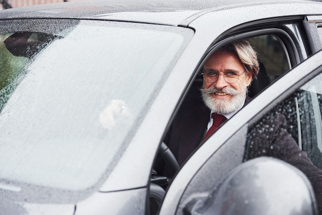 Homem sênior elegante com cabelos grisalhos e barba está ao ar livre na rua, sentado em seu carro.