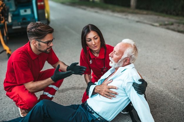 Homem sênior elegante com sintomas de ataque cardíaco, sentado na estrada, trabalhadores de serviços médicos de emergência tentando ajudá-lo. Serviço de assistência ao motorista.