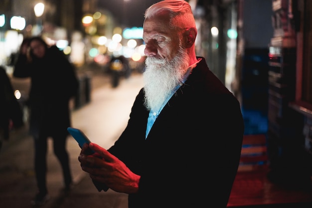 Homem sênior elegante usando smartphone em pé na rua da cidade com luzes de bokeh no fundo - influenciador moderno se divertindo com as tendências da tecnologia - Tecnologia e estilo de vida alegre e idoso - Foco no rosto