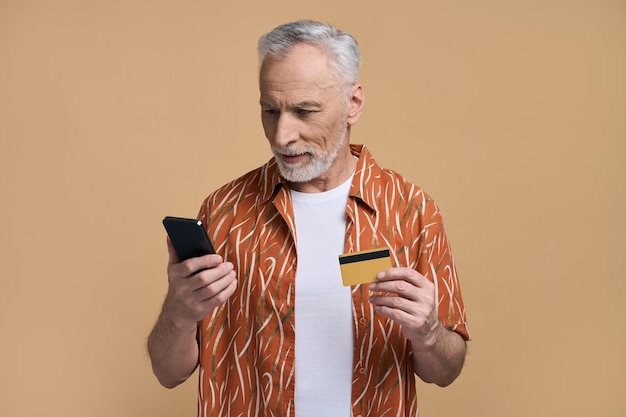 Homem sênior em camisa casual laranja usando telefone móvel inteligente segurando cartão de banco de crédito pano de fundo bege