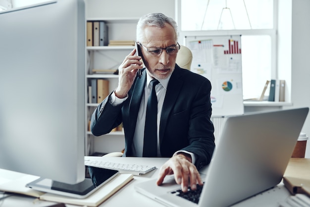 Homem sênior em um elegante terno falando ao telefone enquanto trabalha em um escritório moderno