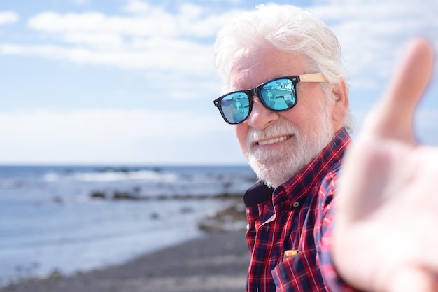 Homem sênior feliz na camisa quadriculada e óculos de sol olhando para a câmera em pé na praia