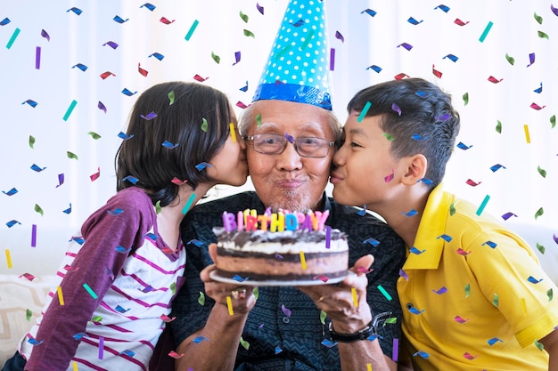 Homem sênior feliz segurando bolo de aniversário e beijado por seus netos com confetes caindo em casa