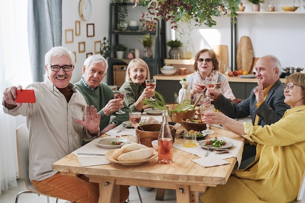 Homem sênior feliz segurando um telefone celular e tirando uma foto com seus velhos amigos durante o jantar à mesa