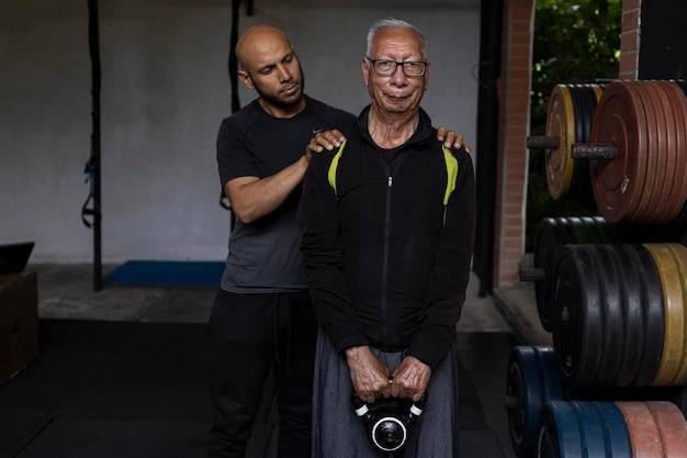 Homem sênior latino-americano com seu personal trainer faz exercícios de reabilitação e terapia segurando um kettlebell com as duas mãos no ginásio Conceito de estilo de vida saudável