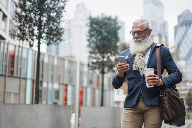 Homem sênior moderno de negócios usando telefone celular enquanto caminhava para o trabalho com a cidade no fundo - rosto de foco