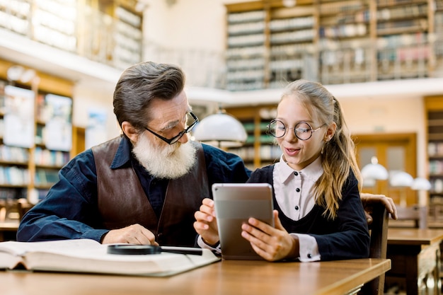 Homem sênior na camisa e colete de couro e neta menina bonita olhando para um tablet, enquanto sentado e estudando juntos na biblioteca. prateleiras de livros antigos no fundo