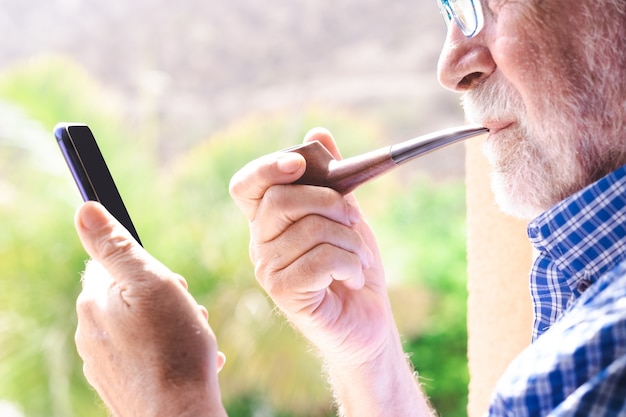 Homem sênior na janela, olhando para o celular, enquanto fuma um cachimbo