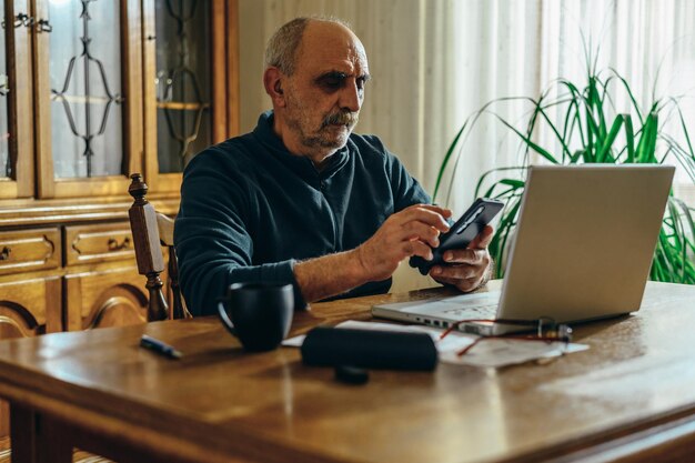 Homem sênior ocupado usando um smartphone e um laptop em casa