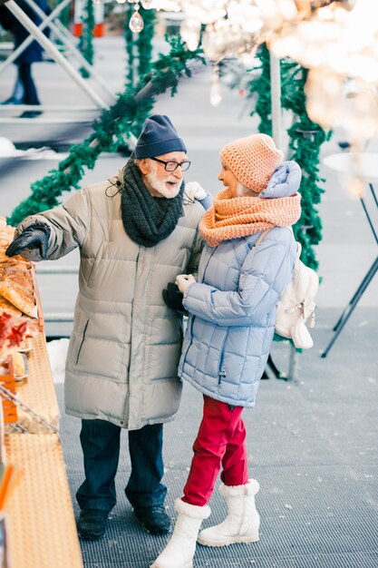 Homem sênior positivo em pé com sua esposa animada no dia de inverno e apontando para a padaria em um balcão na feira