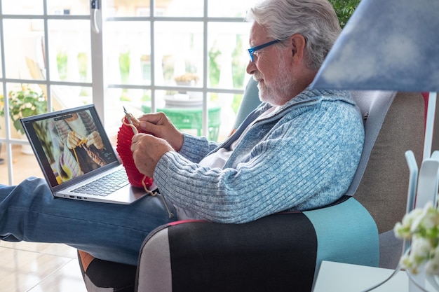 Homem sênior relaxado em casa segue tutorial de tricô on-line para aprender tricô desfrutar de atividade de hobby de lazer sentado na poltrona homens tricotando com ajuda de aula de computador