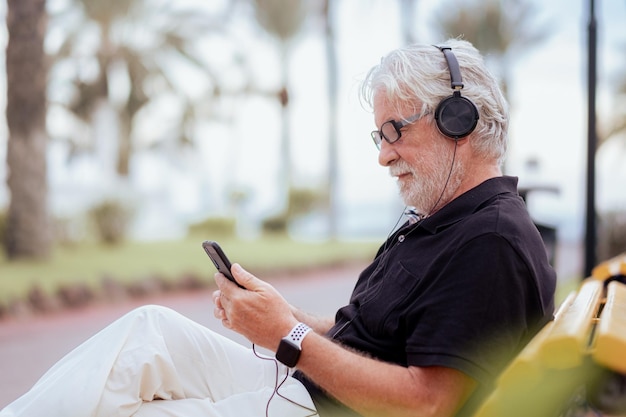 Homem sênior relaxado sentado em um banco amarelo segurando telefone celular usando fones de ouvido Avô idoso barbudo caucasiano no parque público ao ar livre ao pôr do sol