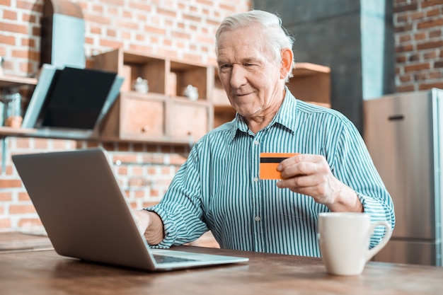 Homem sênior simpático e alegre sorrindo e olhando para a tela do laptop enquanto usa o cartão de crédito