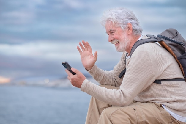 Homem sênior sorridente adulto com mochila descansando durante excursão no mar falando no celular