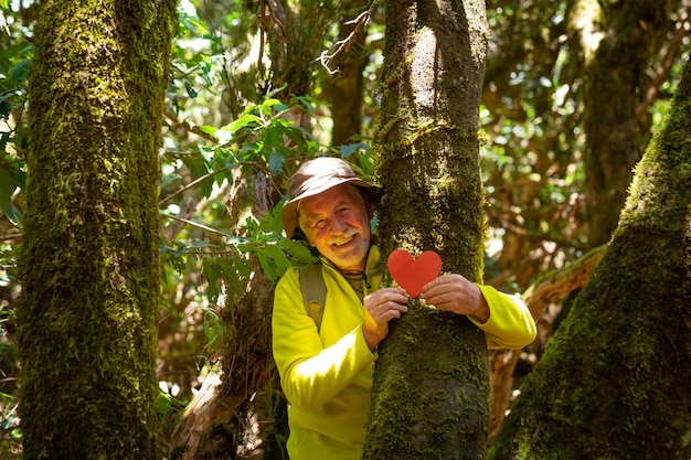 Homem sênior sorridente caminhando na floresta segurando um coração de papel em um tronco de árvore coberto de musgo salvar o planeta do conceito do dia da terra do desmatamento