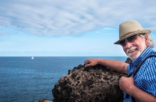 Homem sênior sorridente com chapéu de palha segurando mochila no horizonte de excursão marítima sobre o espaço de cópia de água