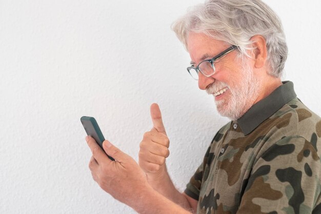 Homem sênior sorridente em pé contra um fundo branco usando telefone inteligente em videochamada com a família