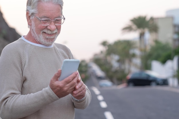 Homem sênior sorridente parado na rua, olhando para o telefone celular, aproveitando a tecnologia e o social