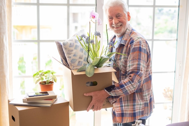 Homem sênior sorridente segurando caixa de papelão posando em casa nova no conceito do dia da mudança de realocação