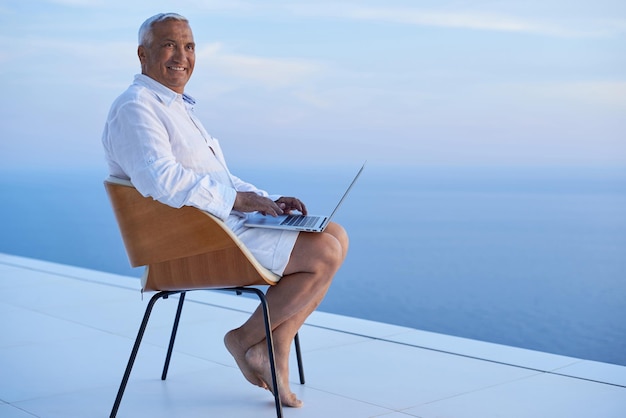 homem sênior trabalhando no computador portátil e olhando o pôr do sol com vista para o mar na varanda da casa moderna