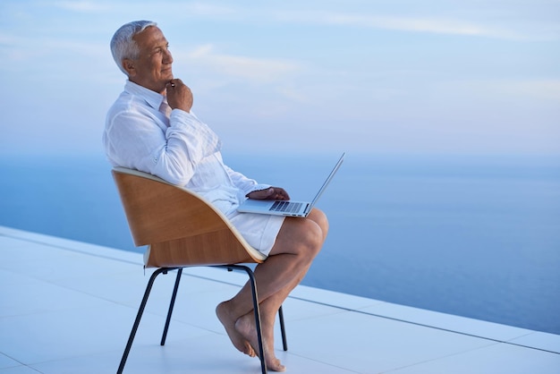 homem sênior trabalhando no computador portátil e olhando o pôr do sol com vista para o mar na varanda da casa moderna