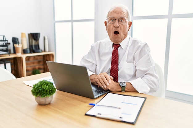 Homem sênior trabalhando no escritório usando laptop de computador com medo e chocado com surpresa e expressão espantada, medo e rosto animado