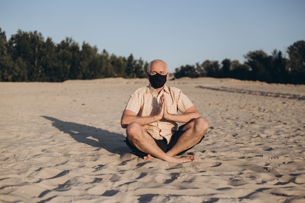 Homem sênior usando máscara protetora de medicina em pose de lótus, sentado na areia. Conceito de calma e meditação.