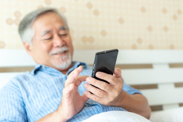 Homem sênior usando um smartphone, sorrindo, sentindo-se feliz na cama em casa - conceito sênior de estilo de vida - selecione o foco e desfoque a fotografia