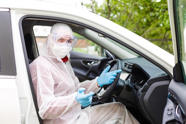 homem senta-se em um carro em traje de proteção, óculos de proteção e uma máscara médica para proteção respiratória