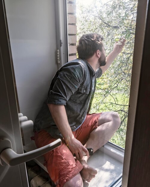 homem senta-se no parapeito da janela e toca e examina raminho de cerejeira no quintal de manhã cedo