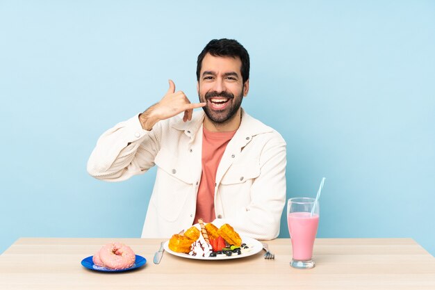 Homem sentado a uma mesa tomando waffles no café da manhã e um milk-shake fazendo gesto de telefone