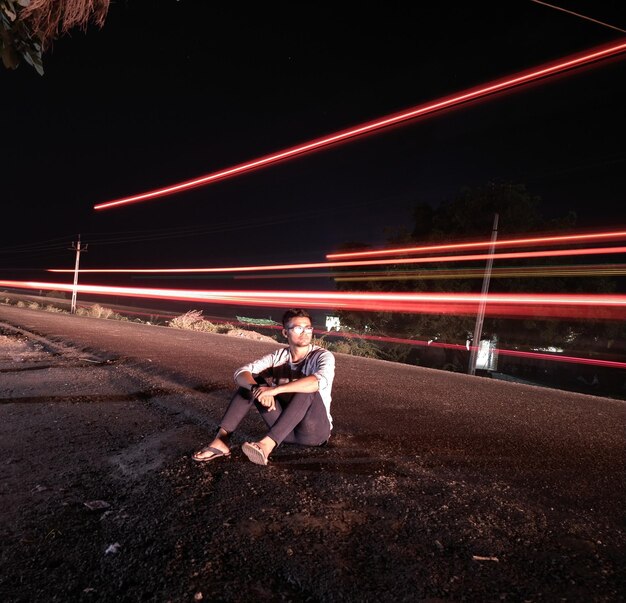 Homem sentado ao lado de uma trilha de luz na estrada à noite