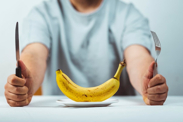 Foto homem sentado atrás de uma mesa com garfo e faca nas mãos e uma banana fresca em um prato