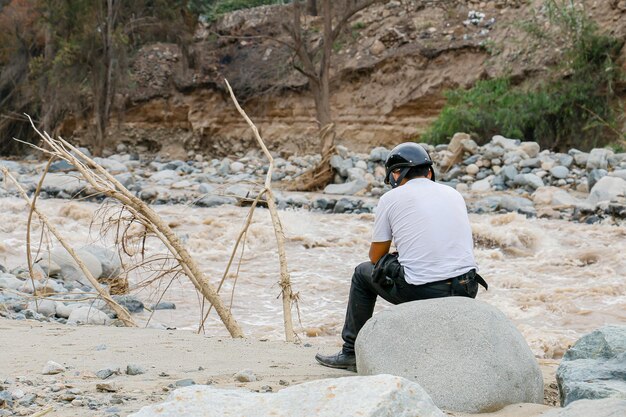 Homem sentado de costas com um capacete na margem de um rio poderoso