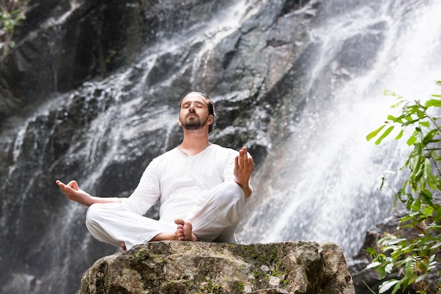 Homem sentado em posição de lótus ioga na rocha sob uma cachoeira tropical e meditando