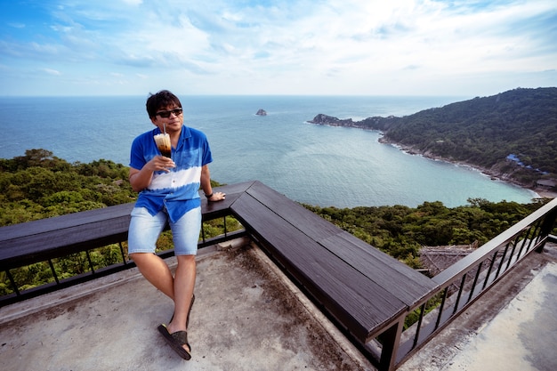 Homem sentado em uma cadeira de madeira no terraço no ponto de vista superior segurando café com leite gelado, homem asiático relaxando com café e vista para o mar