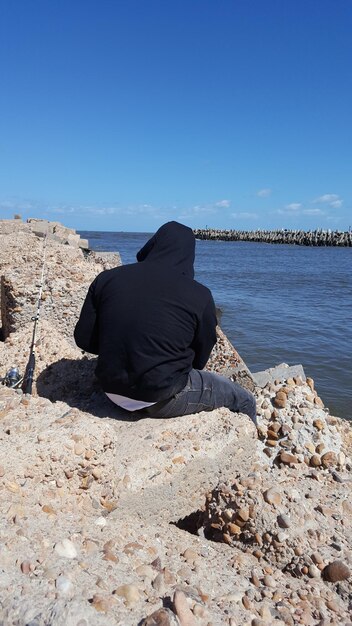 Foto homem sentado em uma rocha na praia contra o céu azul